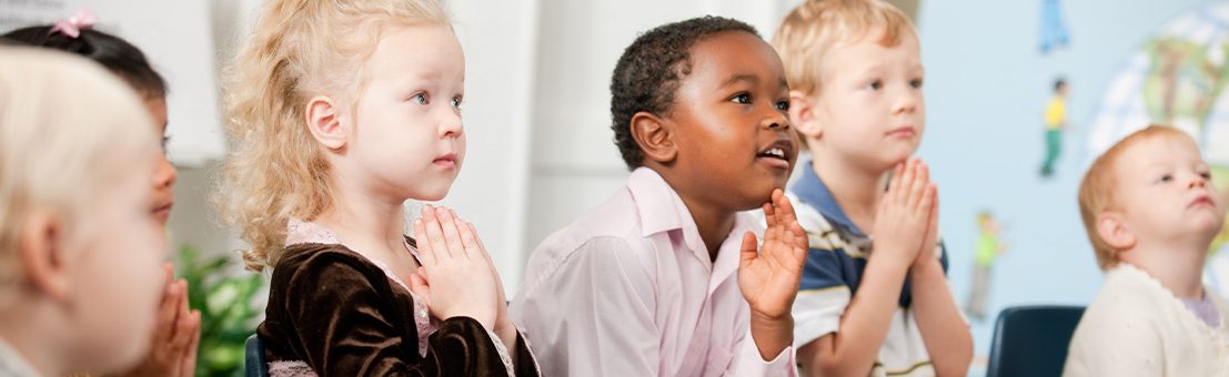 Children with hands in prayer look off camera towards teacher