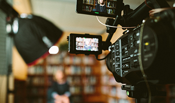 Closeup of video camera in focus, person and bookshelves blurred in background